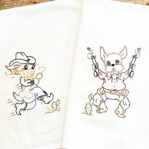 May include: Two white kitchen towels featuring embroidered designs of cartoon animals dressed as cowboys. One animal wears a bandana and hat, while the other holds two pistols. Both designs include cacti and are rendered in brown, blue, and yellow thread.