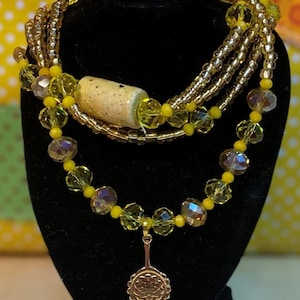 May include: A multi-strand necklace featuring yellow and gold beads, a ceramic bead, and a gold pendant shaped like a fan.