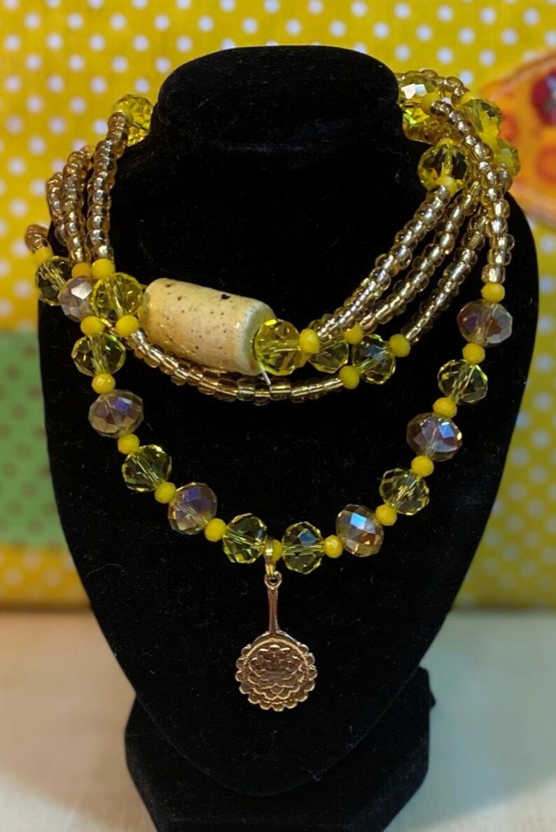 May include: A multi-strand necklace featuring yellow and gold beads, a ceramic bead, and a gold pendant shaped like a fan.