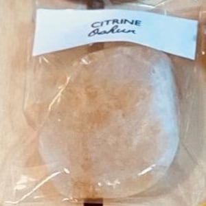 May include: A clear, oval-shaped, translucent, light brown, natural stone with a rough texture. The stone is packaged in a clear plastic bag with a white label that reads "CITRINE Fashion".