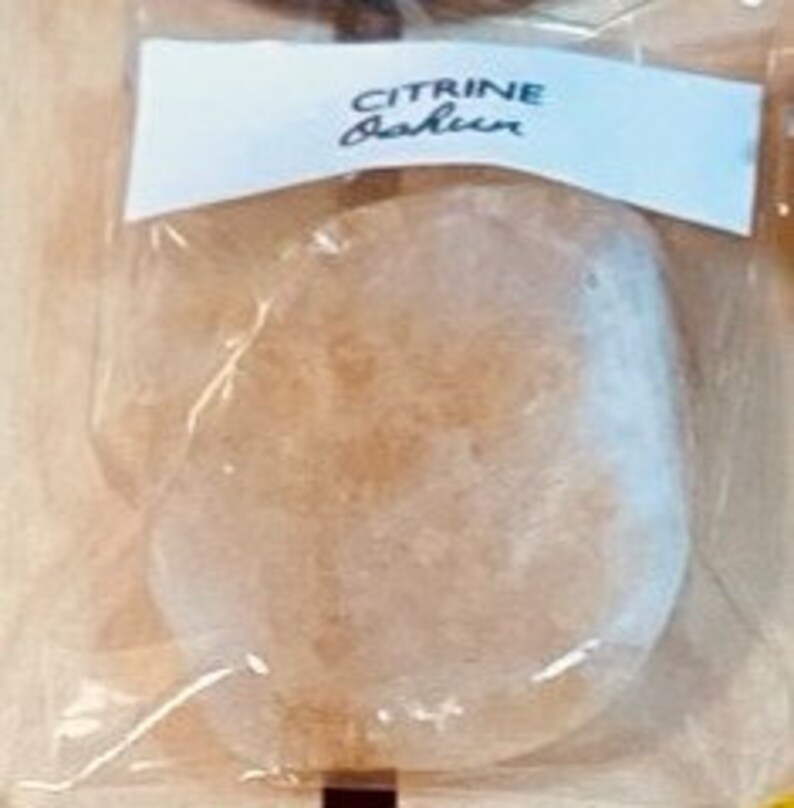 May include: A clear, oval-shaped, translucent, light brown, natural stone with a rough texture. The stone is packaged in a clear plastic bag with a white label that reads "CITRINE Fashion".