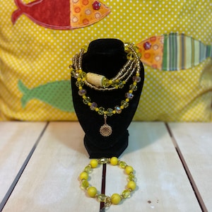 May include: A set of yellow and gold beaded jewellery, including a necklace with a charm and a bracelet with a charm. The necklace is layered with multiple strands of beads and the bracelet has a single strand of beads.