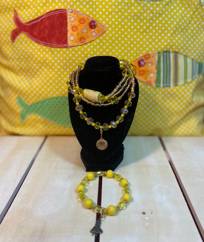 May include: A set of yellow and gold beaded jewellery, including a necklace with a charm and a bracelet with a charm. The necklace is layered with multiple strands of beads and the bracelet has a single strand of beads.