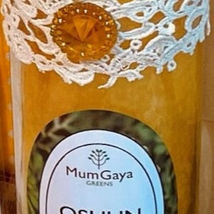 May include: A yellow candle in a glass jar with a white lace ribbon and a gold charm. The label reads "Mum Gaya Greens Oshun Oxum Blessed Candle".