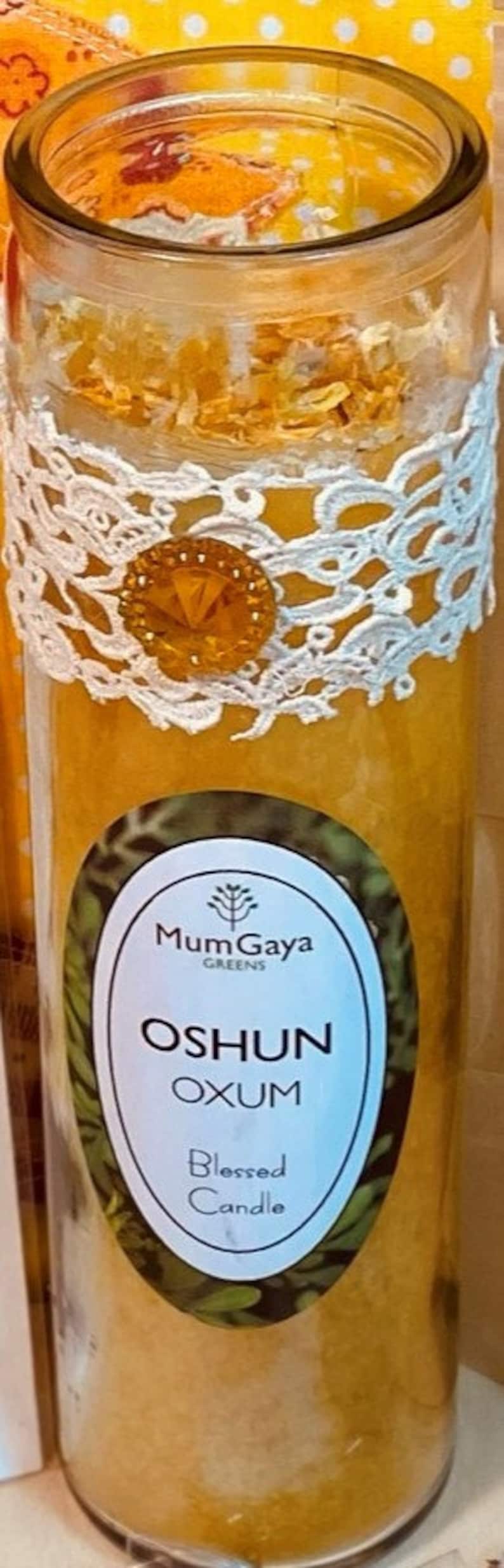 May include: A yellow candle in a glass jar with a white lace ribbon and a gold charm. The label reads "Mum Gaya Greens Oshun Oxum Blessed Candle".