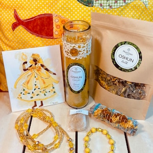 May include: A yellow and gold themed spiritual kit with a candle, bath salts, incense, beads, and a charm. The kit is labelled "Oshun" and features a drawing of a woman in a yellow dress.