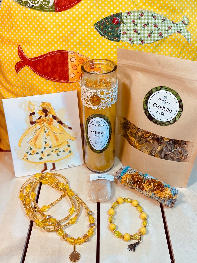 May include: A yellow and gold themed spiritual kit with a candle, bath salts, incense, beads, and a charm. The kit is labelled "Oshun" and features a drawing of a woman in a yellow dress.