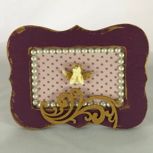 May include: A purple wooden frame with a gold trim and a white pearl border. Inside the frame is a pink and white polka dot background with a gold filigree design and a small white angel figurine with gold wings.