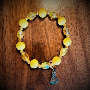 May include: A yellow beaded bracelet with a small charm of a woman in a dress. The beads are a mix of round and faceted.