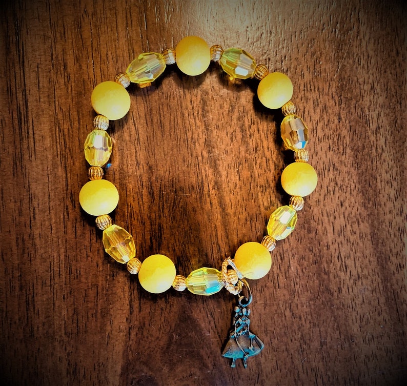 May include: A yellow beaded bracelet with a small charm of a woman in a dress. The beads are a mix of round and faceted.