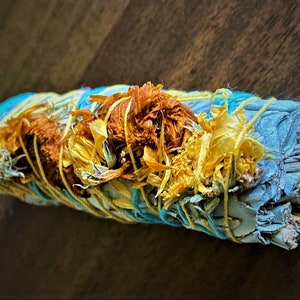 May include: A white sage smudge stick wrapped in blue and yellow string with dried yellow flowers.