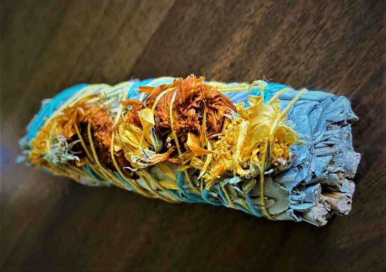 May include: A white sage smudge stick wrapped in blue and yellow string with dried yellow flowers.