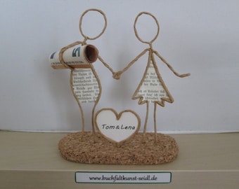 May include: A paper craft wedding figurine of a couple holding hands. The couple is made of brown twine and has a heart-shaped base with the names "Tom & Lena" written on it. The base is made of cork.