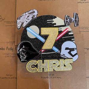 Puede incluir: Un adorno para pastel con temática de Star Wars con un personaje de Darth Vader en blanco y negro, un Stormtrooper blanco, un caza TIE, un Halcón Milenario y un número 7 de purpurina dorada. El nombre "CHRIS" está escrito en letras doradas de purpurina. El adorno para pastel está sobre un fondo marrón.