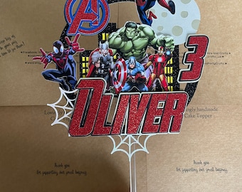 May include: Red, white, and blue Avengers themed cake topper with a red glitter 3 and the name Oliver. The topper features Spiderman, Hulk, Thor, Iron Man, and Captain America.
