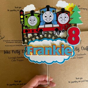 Könnte beinhalten: Ein blauer, roter und grüner Thomas-die-Lokomotive-Kuchentopper mit einer weißen Wolke und dem Namen Frankie in blauen und roten Buchstaben. Die Zahl 8 ist in rot und gelb. Der Topper ist auf einem durchsichtigen Plastikstab.