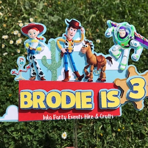 Pode incluir: Um topo de bolo colorido com personagens da franquia de filmes Toy Story. O topo inclui Woody, Jessie, Buzz Lightyear, Bullseye e um cacto. O topo também inclui o texto "Brodie is 3" e o logotipo da empresa "Inka Party Events Hire & Crafts".