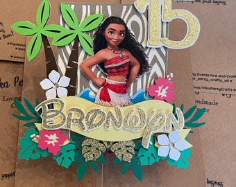 Pode incluir: Um topo de bolo com uma personagem de Moana com um 15 dourado, uma faixa amarela com o nome "Brondyn" em letras douradas com glitter e folhagem tropical.
