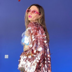 May include: A rose gold sequin jacket with long fringe. The jacket is worn over a sequined top and blue shorts. The background features a blue wall, a pink ceiling, and a colorful chandelier.