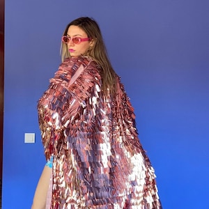 May include: A long, shimmering, rose gold sequin coat. The coat is the focal point, reflecting light. The background features a blue wall, a pink ceiling, and a colorful chandelier. The overall aesthetic is glamorous and bold.