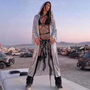 May include: A long, hooded, silver sequinned robe worn over a black harness-style top and black sheer skirt. The garment features wide sleeves and a shimmering, reflective surface that catches the light, creating a metallic appearance.