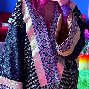 May include: A black sequined kimono-style jacket featuring wide sleeves and a prominent front border with a colorful geometric pattern in shades of blue, pink, and gold. The garment has a metallic gold trim and a tan interior lining visible at the cuffs.