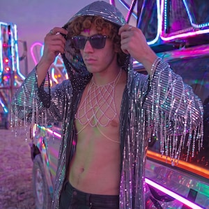 May include: A long, hooded silver sequin duster coat featuring long fringe detailing on the sleeves, paired with a delicate silver-toned chain body harness worn over the chest, and dark-coloured trousers with black sunglasses.