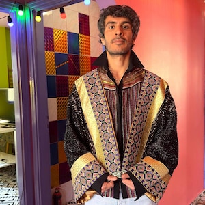 May include: A long-sleeved jacket featuring black fabric with a shimmering texture, accented by wide gold bands and intricate geometric patterns in purple, blue, and orange. The garment has a high collar, wide cuffs, and a structured open front design.