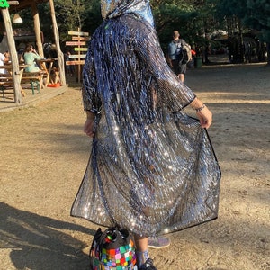 Puede incluir: Una chaqueta con capucha de lentejuelas plateadas con un diseño largo y fluido. La chaqueta se lleva sobre una camisa negra y la persona lleva zapatillas azules. La persona está de pie en un camino de tierra con una bolsa colorida a sus pies.