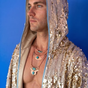 Puede incluir: Una bata con capucha cubierta de lentejuelas doradas brillantes con un borde azul claro. La bata se lleva con tres collares de cobre superpuestos, cada uno con una piedra turquesa central, incluyendo un colgante en forma de media luna.