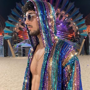 Puede incluir: Una bata con capucha cubierta de rayas verticales de lentejuelas brillantes en tonos azul, morado, verde azulado y dorado. La prenda presenta un diseño largo abierto por delante con capucha, reflejando la luz sobre su superficie metálica.