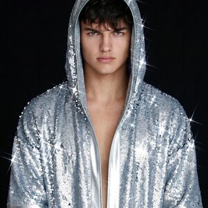 May include: A short-sleeved hooded robe covered in shimmering silver sequins. The garment features a smooth, metallic silver trim along the front opening and the sleeve cuffs, creating a reflective and high-shine appearance throughout the piece.
