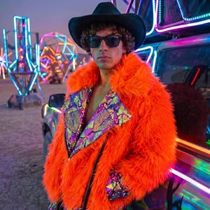 May include: A bright orange faux fur jacket featuring a black zipper closure and wide lapels adorned with iridescent purple and green sequins. The jacket is worn with a black cowboy hat and dark sunglasses, creating a bold, textured appearance.
