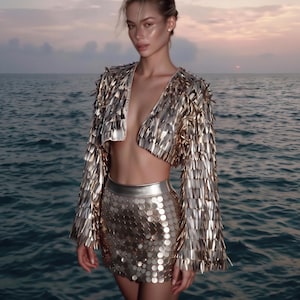 May include: A gold cropped jacket with long flared sleeves covered in metallic fringe and a matching high-waisted mini skirt featuring a dense pattern of circular gold sequins. The set has a reflective, shiny finish throughout the garment.