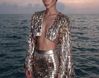 May include: A gold cropped jacket with long flared sleeves covered in metallic fringe and a matching high-waisted mini skirt featuring a dense pattern of circular gold sequins. The set has a reflective, shiny finish throughout the garment.
