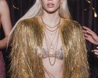 May include: A gold fringe jacket featuring long metallic strands, worn over a silver rhinestone-embellished bra and matching bottoms. The outfit is accessorized with a gold halo headpiece, long dangling crystal earrings, and layered gold chain necklaces.