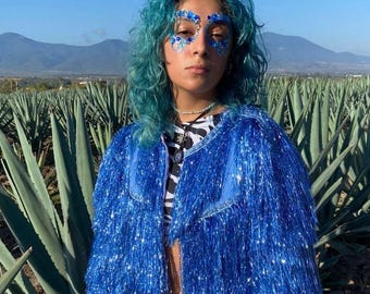 Puede incluir: Una chaqueta azul brillante cubierta con largas capas de flecos metálicos relucientes. La chaqueta se lleva sobre una camiseta con estampado de cebra en blanco y negro y vaqueros de mezclilla claros. Se completa con brillos azules en los ojos.