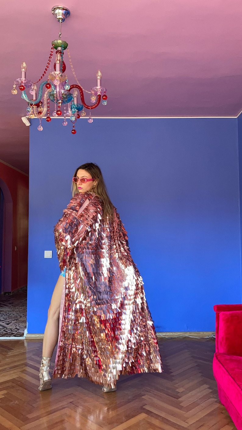 May include: A long, shimmering, rose gold sequin coat. The coat is the focal point, reflecting light. The background features a blue wall, a pink ceiling, and a colorful chandelier. The overall aesthetic is glamorous and bold.