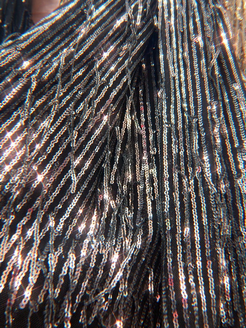 May include: A black fabric garment featuring vertical rows of shimmering silver sequins. The material is adorned with long, dangling fringe strands covered in reflective sequins that catch the light, creating a dense and textured appearance.