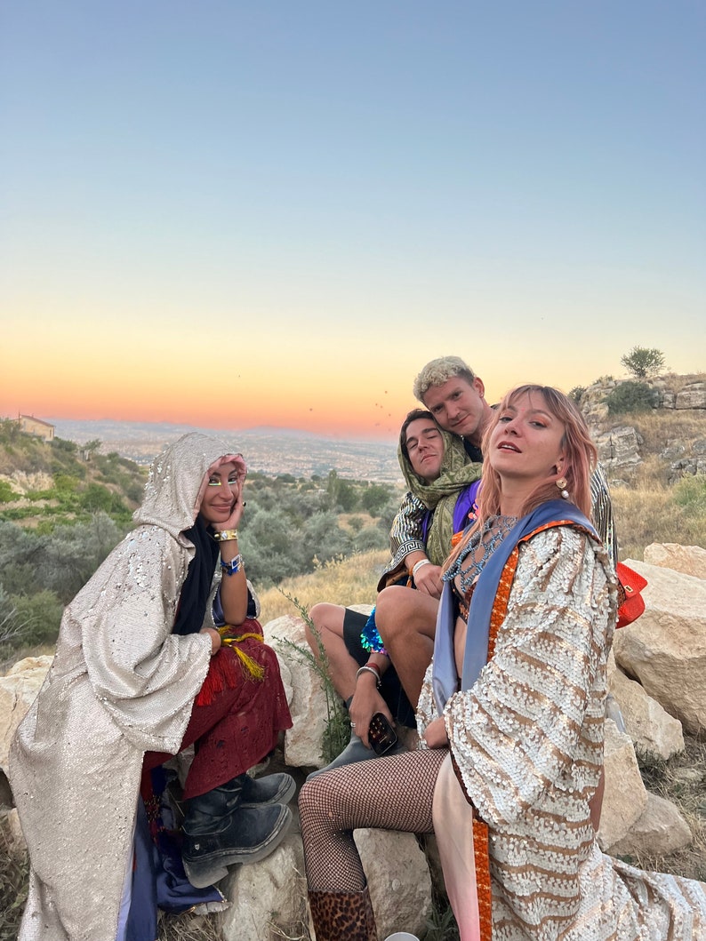 May include: A group of people wearing sequined hooded cloaks, patterned robes, fishnet stockings, and leather boots. The garments feature metallic gold and silver sequins, intricate embroidery, and layered fabrics in shades of red, blue, and tan.