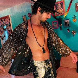 May include: A black fedora hat, a long-sleeved open-front jacket covered in gold sequins, and black trousers with a gold and white celestial print. A long black beaded necklace with a tassel hangs around the neck over the bare chest.