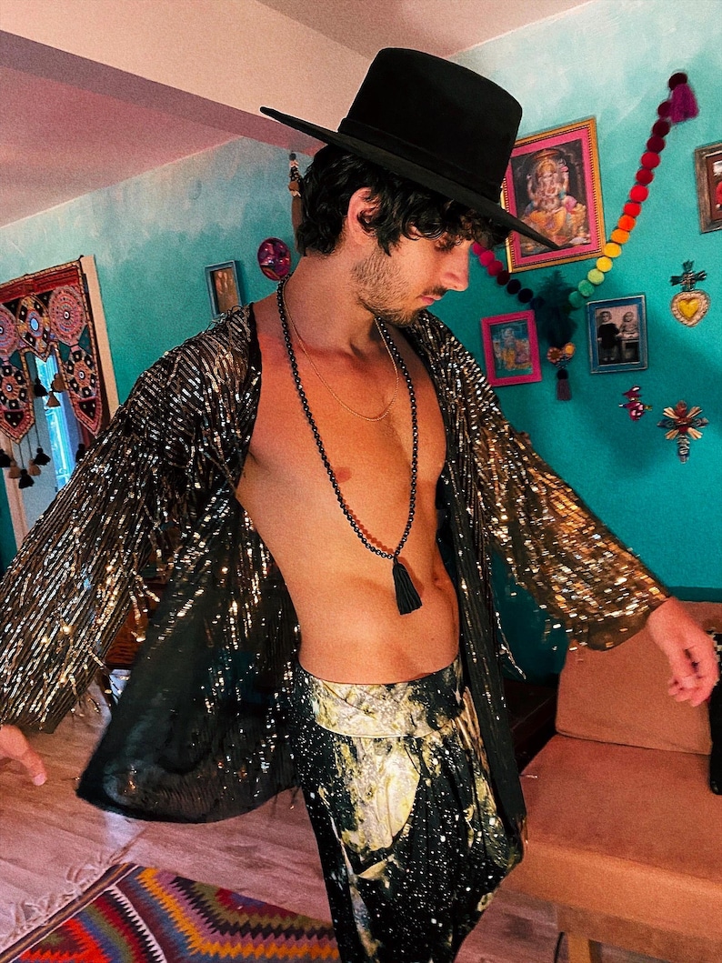 May include: A black fedora hat, a long-sleeved open-front jacket covered in gold sequins, and black trousers with a gold and white celestial print. A long black beaded necklace with a tassel hangs around the neck over the bare chest.
