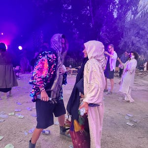 May include: Two individuals wearing hooded cloaks, one featuring vibrant multicolored sequins and the other in a shimmering gold fabric. Both outfits include dark shorts, layered garments, and sturdy boots, captured in an outdoor setting with purple lighting.