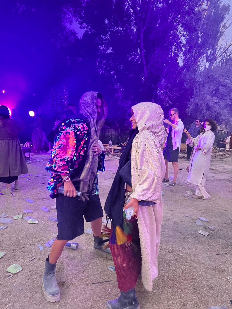 May include: Two individuals wearing hooded cloaks, one featuring vibrant multicolored sequins and the other in a shimmering gold fabric. Both outfits include dark shorts, layered garments, and sturdy boots, captured in an outdoor setting with purple lighting.