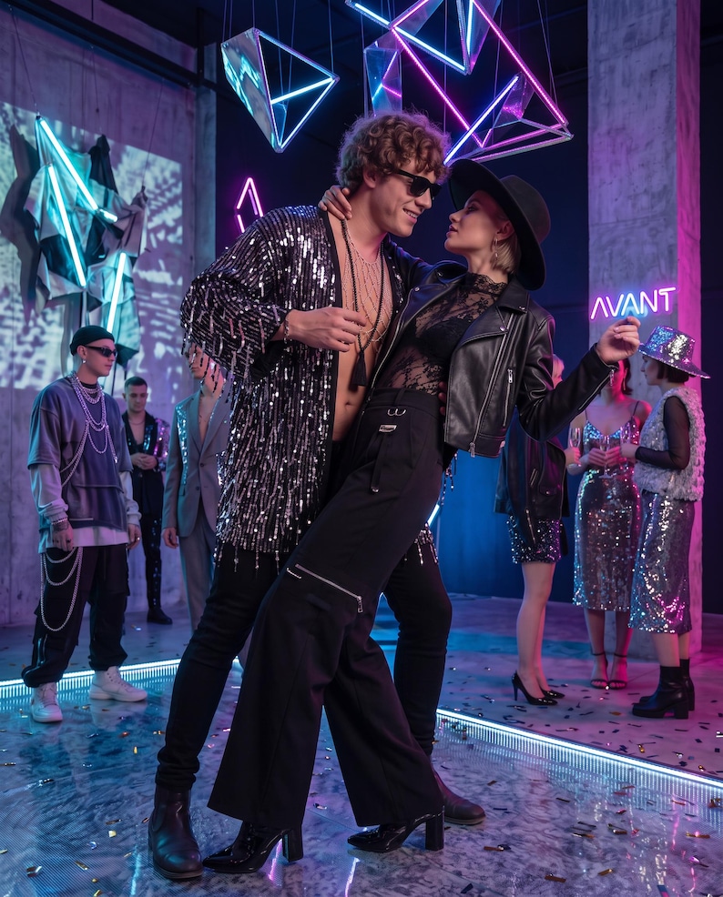 May include: A black sequined open-front jacket with fringe, black trousers with zipper details, a black leather jacket, a black lace top, a black wide-brimmed hat, and black sunglasses. The scene includes neon lighting and the text AVANT.