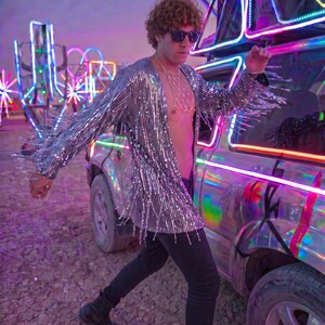 May include: A long-sleeved silver sequined jacket featuring long fringe detailing along the sleeves and hem. The jacket is worn open over a gold-toned chain body harness, paired with black trousers and black boots, with dark sunglasses.