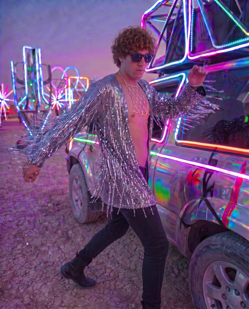 May include: A long-sleeved silver sequined jacket featuring long fringe detailing along the sleeves and hem. The jacket is worn open over a gold-toned chain body harness, paired with black trousers and black boots, with dark sunglasses.