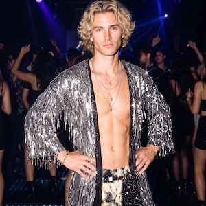 May include: A silver sequined open-front cardigan with long sleeves and fringe detailing, worn over dark trousers with a celestial print. The garment is paired with a layered silver chain necklace featuring small pendants and a beaded bracelet.