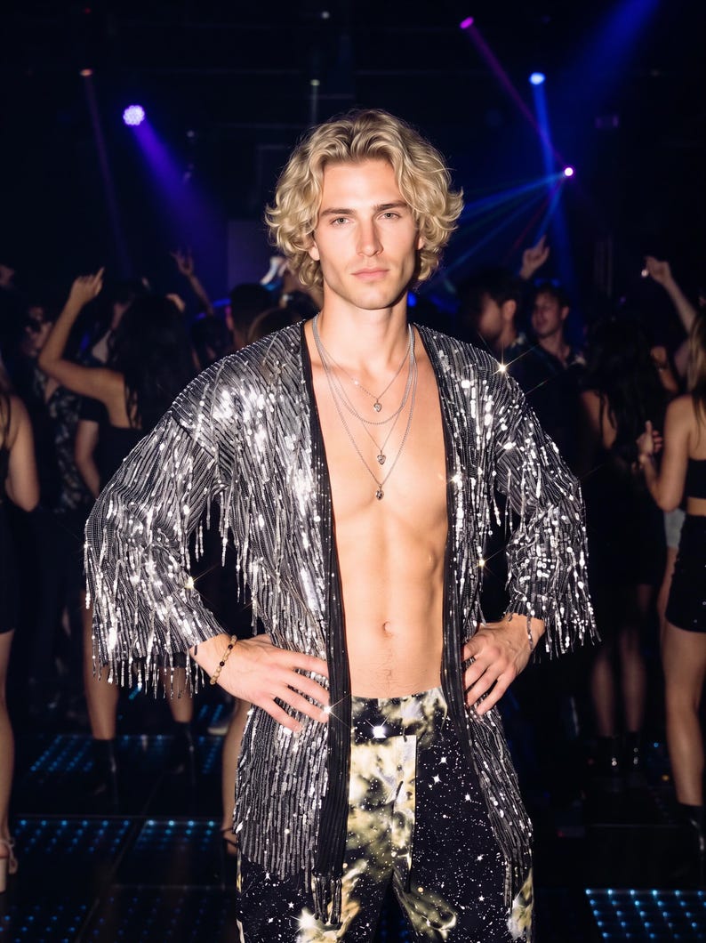 May include: A silver sequined open-front cardigan with long sleeves and fringe detailing, worn over dark trousers with a celestial print. The garment is paired with a layered silver chain necklace featuring small pendants and a beaded bracelet.