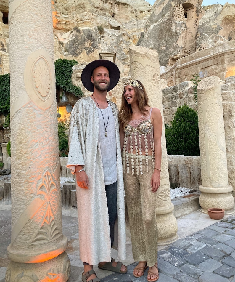 May include: A person wearing a wide-brimmed dark hat, a long shimmering silver duster, a light gray shirt, and dark pants, standing next to a person wearing a gold embellished crop top with hanging floral accents, gold pants, and a floral headpiece.